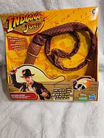 Front view of the retail box showing Indiana Jones cartoon, whip coiled inside visible through clear plastic, and brand logos.