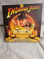 Front of the Indiana Jones: Sands of Adventure board game box, showing the game's title and artwork featuring Indiana Jones and a woman with desert motifs including snakes and ancient symbols.