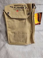 Front view of beige canvas satchel bag with black print 'Property of Dr. H. Jones Jr.' and snap button flap closure, showing attached tag.