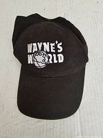 Front view of black baseball cap showing white embroidered 'WAYNE'S WORLD' text and globe logo centered on front panel.