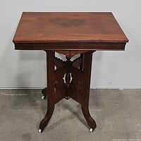 Front view of antique Eastlake style pedestal table, showing square top and carved base with castered feet.