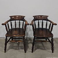 Pair of matching antique wooden captain's chairs shown together on concrete floor against white wall.