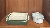 Green rectangular ceramic baker with white interior and handles alongside a white round ceramic baker with clear glass lid, displayed on wood shelf.