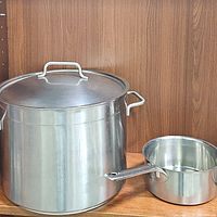 Large stainless steel stock pot with lid and smaller pot placed side by side on a wooden shelf.