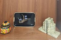 Overview photo showing jarred lacquer container, black lacquer serving tray with floral inlay, and pair of marble bookends on wooden shelf.