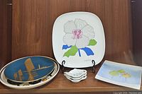 Group photo showing multiple decorative plates: blue and gold round plate, large white flower square plate, small tea bag holder dishes stacked, and yellow ginkgo leaf plate