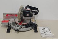 Front view of the Delta Sidekick 12" compound miter saw showing sliding fence, blade guard, and control handle with cord, plus included instruction manual.