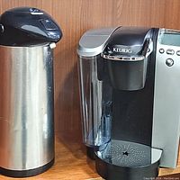 Keurig coffee maker with visible water reservoir and controls next to the stainless steel coffee carafe