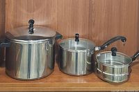 Three vintage Farberware pots displayed side by side on a wooden shelf - large stock pot with lid, medium saucepan with handle and lid, two smaller stackable pots with lids