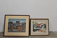 Two framed pictures leaning against a wall on concrete floor. The left is a colorful cityscape print with chairs and trees by Michel Delacroix; the right is a framed watercolor of a covered bridge.