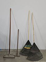 Four rakes standing against a wall: two leaf rakes with metal black tine heads and wooden handles on the right, two bow rakes with straight metal tine heads and wooden handles on the left.
