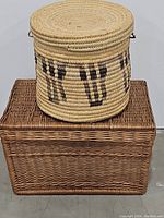 Round woven basket with handles and geometric black pattern on sides seated atop rectangular wicker basket trunk with hinged lid.