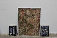 Three framed art pieces displayed against a white wall on the floor: large central silver-framed abstract portrait flanked by two smaller framed paintings on each side.