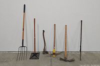 Full view of the six garden and digging tools against a white wall and concrete floor, showing wooden handles and metal working heads.