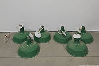 Six green vintage industrial pendant lamps placed on the floor, showing top and side views of the lamps with attached white canopy covers and visible wiring.