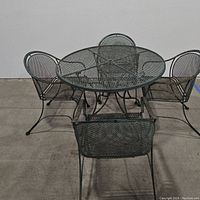 Top angled view of the wrought iron patio set including the table and four matching chairs on a concrete floor against a wall.