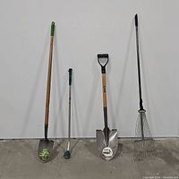 Full view of two shovels, one hose extension, and one metal leaf rake arranged side by side against white wall and concrete floor.
