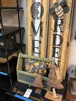 Four bee-themed painted wood signs displayed vertically with text and honeycomb designs, along with green wood and metal carrier holding cloth pillows and wooden bee decorations on a black shelf.
