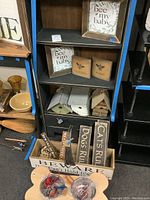 Shelving unit with wood plaques featuring bee themes, multiple small wood birdhouses painted white and natural wood, pet signs with inscriptions, and a wooden crate with assorted wood decor pieces in front.