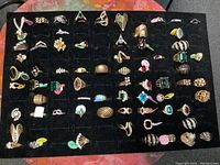 Overview of approximately 69 costume rings arranged on a black foam display pad in rows, showing a variety of colors, designs, and shapes.