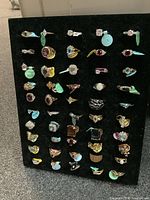 Black foam display pad with approximately 50 assorted costume rings mounted in rows, many with colored size tags