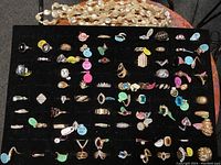 Black foam display pad holding assortment of rings with colored size tags and multi-strand faux pearl necklace draped above