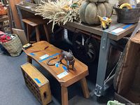 Front view of the industrial work table showing wooden top and metal legs, with various other furniture items and objects nearby. Table wheels are visible at the bottom of the metal legs.