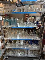 Full view of the 6-tier metal shelving unit loaded with various glassware, bottles, and decorative items. Shelves are made of wire grid metal with chrome finish.