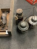 Three vintage railroad lanterns with colored glass globes and metal wire guards, showing signs of rust and scratches, placed on carpeted floor near a wood box with shoe forms.