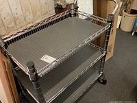 Side and angled view of stainless steel rolling cart with plastic liners on shelves. Black caster wheels visible at base.