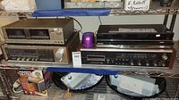 Four vintage stereo components on metal shelving: Realistic dual cassette player, Realistic AM/FM 8-track stereo receiver, Sony BluRay player, and Samsung BluRay player. Purple LED or lamp accessory also present.