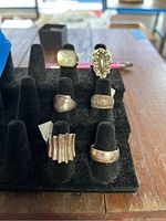 Six sterling silver rings on black ring display stand, various designs including ridged, engraved floral, filigree, plain dome, star-engraved, and inlaid stone designs, showing tarnish and scratches.