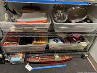 Two-tier wire shelf holding plastic storage bins with office supplies, cookware, sewing notions and model Texaco ship on floor