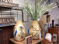 Photo showing two beige pottery vases with black tree branch designs placed on a wooden surface, along with a wooden word art sign reading 'Love never fails.'