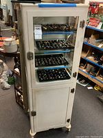 Front view of painted metal cabinet showing glass door, interior glass shelves with contents, and single key in lock