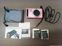 Pink 4K 44MP digital camera with accessories spread out including two batteries, USB cable, soft pouch, and instruction manual.