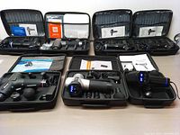 Seven handheld muscle massage tools shown in their carrying cases containing the massage guns, multiple attachments, manuals, and power cords.