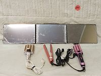 Photo showing all four items: LED triple mirror and three hair styling tools (beige and pink triple barrel curling irons, red hair straightener) placed on beige carpet.