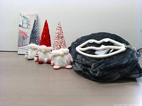 Photo showing three mini Santa Claus decorations with different colored hats, a dark grey heated blanket folded, and a white lips-shaped decorative lamp next to them.