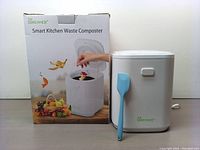 Photo of the Qmcahee Smart Kitchen Waste Composter box and the compost bin unit with blue spatula placed on the side. Shows the product branding and images on the box.