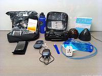 Overview of bundled items on table including heated jacket in plastic bag, massage gun, hydration bladder, projector light, power adapter, portable fan, blue water bottle, and other electronics and accessories.