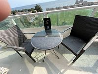Photo showing two black woven rattan-style chairs on either side of a round glass-top bistro table with umbrella hole on a balcony with ocean and park view.