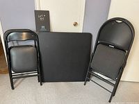 Image showing black plastic square card table with four black folding chairs, two larger with upholstered seats and two smaller with faux leather seats.