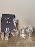 Overall view of vintage crystal decanter with stopper, four crystal tumblers, and four liqueur glasses on table with lot number sign.