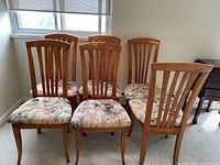 View of all six wooden dining chairs arranged side by side in a room with carpet and a window in the background.