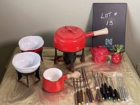 Photo of vintage red enamel fondue pot with wooden handle on black metal stand with wooden feet and burner beneath, two white ceramic bowls on black stands, red mini covered pot, red small saucepan with wooden handle, set of fondue forks with varied colored tips and handles arranged on table.