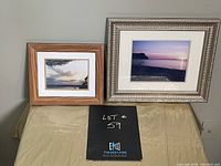 Two framed photographs of lake scape in two different frames on a cloth-covered table with a sign with lot number.