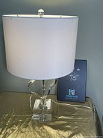 Full view of the clear acrylic stem lamp with white drum shade on a gold surface.