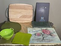 Photo showing the full collection of seven kitchen items: olive green lazy susan, wooden cutting board with metal handles, large glass trivet with pink floral and vintage text design, green plastic colander, green silicone pot holder, rectangular dark stone cheese board, and metal cheese fork with multiple prongs.