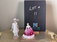 Photo showing both porcelain figurines with black box in background marked 'LOT #11'.